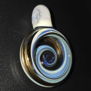 May include: A glass pendant with a swirling spiral design. The pendant features a combination of blue, white, and brown colors. The top loop is white. The spiral design is centered in the pendant.