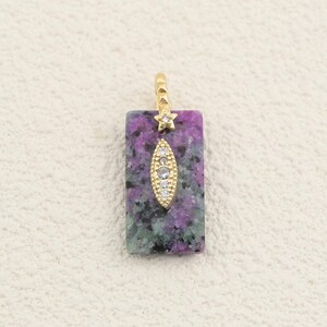 May include: A rectangular pendant featuring a mottled purple and green stone base. A gold-toned metal bail sits at the top, connected to a small, marquise-shaped gold-toned accent piece embedded with tiny clear stones centred on the stone surface.