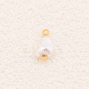 May include: A single, white, teardrop-shaped pearl pendant with a gold-colored bail and base. The pearl has a slightly irregular surface. The pendant is set against a neutral, textured background.