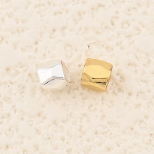 May include: Two small, geometric beads. One is silver-colored, the other is gold-colored. The beads have a faceted design, catching the light. They are likely components for jewelry making or crafting.