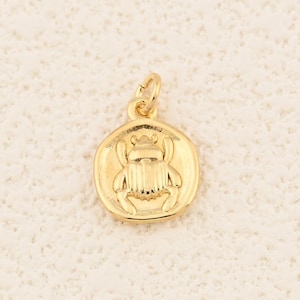 May include: A gold-colored pendant with a detailed scarab beetle design. The pendant is round with a small loop at the top for a chain. The beetle is centered within a circular frame, showcasing intricate details.