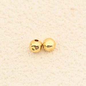 May include: Two small, round, gold-colored beads. One bead has a hole, and the other is solid. The beads are likely for jewelry making or crafting. The beads are on a textured, off-white surface.