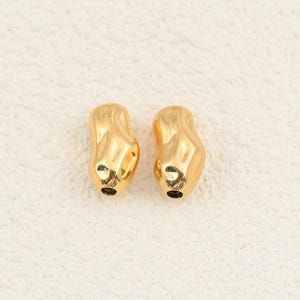 Può includere: Due piccole perline color oro dalla forma irregolare, organica e leggermente allungata. Ogni perla presenta una superficie metallica liscia e riflettente con un foro centrale visibile alla base, caratterizzata da una finitura lucida.