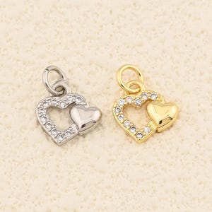 May include: Two heart-shaped pendants. One is silver-toned with clear crystals around the outer heart and a solid heart inside. The other is gold-toned with clear crystals around the outer heart and a solid gold heart inside. Both have small loops for hanging.