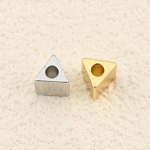 May include: Two small, triangular metal objects. One is silver-colored, the other is gold-colored. Each has a circular hole in the center. The objects are likely small components or decorative elements.