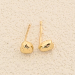 May include: A pair of gold-colored stud earrings. Each earring has a rounded, irregular shape, with a post for insertion into the ear. The earrings are displayed on a cream-colored surface.
