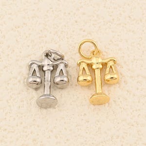 May include: Two small scale charms, one silver-colored and one gold-colored, are displayed. The charms are detailed with a balance beam design, featuring two hanging pans. The gold charm has a loop for attachment. These charms are suitable for jewelry making.