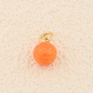 May include: A small, round orange charm with a textured surface and a gold-colored loop. The charm is suspended against a neutral background, highlighting its vibrant color and simple design. This jewelry piece could be a pendant or a bead.