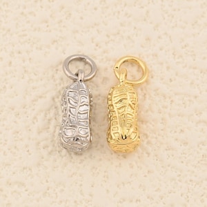 May include: Two peanut-shaped charms, one silver and one gold, each with a small loop at the top. The charms have detailed textures, resembling the surface of a peanut. They are likely intended for jewellery making or as decorative elements.