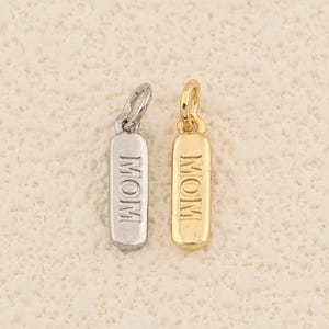 May include: Two rectangular pendants, one silver-toned and one gold-toned, each engraved with the word "MOM". The pendants have a small loop at the top for attaching to a chain or bracelet. The background is a light beige color.