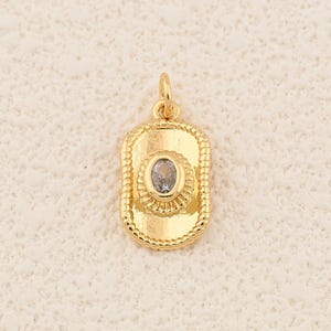 May include: A gold-colored pendant with an oval-shaped, light-colored stone in the center. The pendant has a rectangular shape with a decorative border and a small loop at the top for a chain. The pendant is approximately 0.75 inches long.