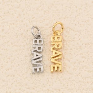 May include: Two metal pendants, one silver and one gold, each inscribed with the word "BRAVE" in capital letters. The pendants are vertically aligned and feature a small loop at the top for attachment. The background is a textured, light beige surface.