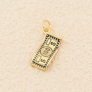 May include: A gold-coloured charm shaped like a £100 note. The charm has a small loop at the top for attaching to a necklace or bracelet. The note design includes the number 100 and dollar signs.