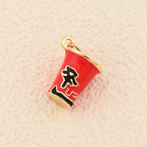 Puede incluir: Un charm rojo y dorado con forma de taza de ramen. El charm tiene texto negro en la parte delantera y un borde dorado. El charm probablemente está hecho de metal y está diseñado para ser un colgante o charm.