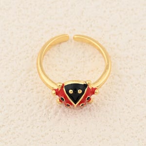 May include: A gold-colored adjustable ring featuring a ladybug design. The ladybug has a black head and a red body with black spots. The ring is open at the bottom for size adjustment.