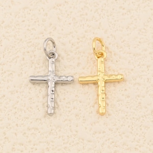 May include: Two small cross pendants. One is silver-colored, the other is gold-colored. Both crosses have a textured design and a small loop at the top for attaching to a chain or other jewelry. The background is a light beige color.