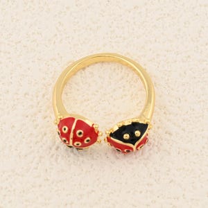 Pode incluir: Anel dourado com dois desenhos de joaninhas, um vermelho e outro preto, com pontos detalhados. O anel tem um design aberto, permitindo o ajuste do tamanho. Os elementos da joaninha estão posicionados em ambos os lados da abertura do anel.