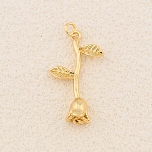 May include: A gold-coloured rose pendant with a stem and two leaves. The rose is in full bloom, and the leaves have detailed veining. The pendant has a loop at the top for attaching to a chain or other jewellery.