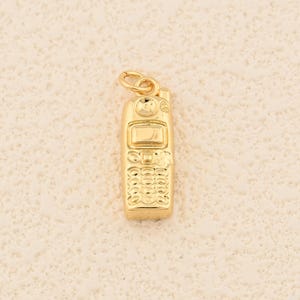 May include: A gold-colored charm in the shape of a vintage cell phone. The charm has a rectangular body with a screen, keypad, and a small loop at the top for attaching to a necklace or bracelet. The surface has a textured appearance.