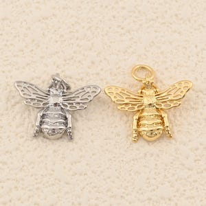 May include: Two bee-shaped charms. One is silver-colored, and the other is gold-colored. Both charms have detailed wings and body segments, with a loop at the top for attaching to a chain or other jewelry. The background is a light beige color.