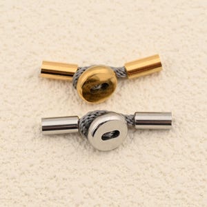 May include: Two toggle clasps, one gold-toned and one silver-toned, each with a button-shaped center and cylindrical ends. The clasps are connected by a gray cord, suitable for jewelry or craft projects.