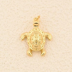May include: A gold-colored turtle charm with detailed texture on its shell, flippers, and head. The charm has a loop at the top for attaching to a necklace or bracelet. The turtle is a symbol of longevity and wisdom.