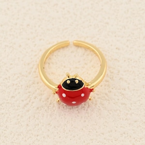 Könnte beinhalten: Verstellbarer goldfarbener Ring mit einem roten Marienkäfer-Design mit weißen Punkten und einem schwarzen Kopf. Der Ring ist vor einem neutralen Hintergrund platziert, der die leuchtenden Farben des Insekts hervorhebt.