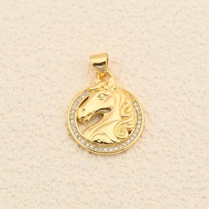 May include: A gold-colored pendant featuring a horse head profile within a circular frame. The horse head is detailed with flowing mane and facial features. The frame is encrusted with small, clear gemstones, adding sparkle. The pendant has a loop for a chain.