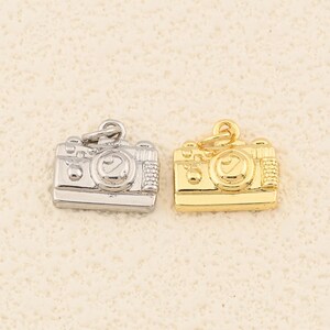 May include: Two small, detailed camera charms. One is silver-colored, the other is gold-colored. Both charms have a vintage camera design with intricate details, including a lens and buttons. The charms are likely for jewelry making or crafting.