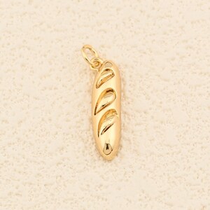 Könnte beinhalten: Ein goldfarbener Anhänger in Form eines Baguettes. Der Anhänger hat drei diagonale Schnitte auf der Oberseite und eine kleine Schlaufe zum Befestigen an einer Kette. Das Baguette hat eine glänzende Goldfarbe.