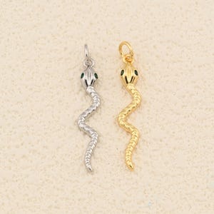 May include: Two snake-shaped pendants. One is silver with green eyes, the other is gold with black eyes. Each has a small loop at the top for hanging. The pendants are displayed against a neutral background.