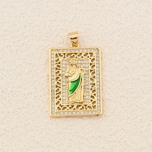 Puede incluir: Colgante rectangular dorado que representa una figura religiosa con una prenda verde. El colgante tiene un borde decorativo y está incrustado con pequeños cristales transparentes. Un pequeño lazo en la parte superior para una cadena.
