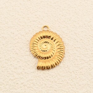 以下が含まれることがあります： 黄金色のオウムガイの貝殻の形をしたペンダント。ペンダントは、詳細な隆起とチェーンまたはコードに取り付けるための上部の小さなループを備えたスパイラルデザインです。背景は、テクスチャ加工されたオフホワイトの表面です。
