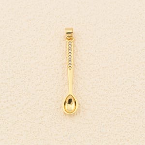 May include: A gold-coloured spoon pendant with a small, oval bowl and a handle decorated with small, clear gemstones. The pendant is designed to be worn as jewellery.