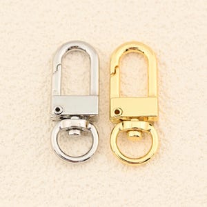 May include: Two metal swivel clasps, one silver and one gold, are displayed side-by-side. Each clasp features a rectangular base, a D-shaped swivel, and a circular ring. The clasps are designed for attaching to bags, keychains, or other accessories.