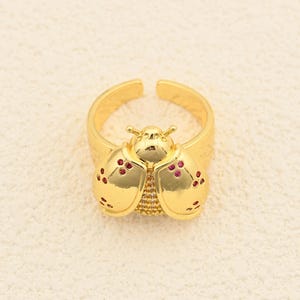 May include: A gold-coloured adjustable ring with a ladybug design. The ladybug has red accents on its wings and a textured body. The ring is set against a neutral background.