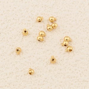 Könnte beinhalten: Eine Ansammlung kleiner, glänzender, goldfarbener Perlen. Jede Perle ist kugelförmig mit einem kleinen Loch. Die Perlen sind auf einer hellen Oberfläche verstreut, was darauf hindeutet, dass sie lose Komponenten für die Schmuckherstellung oder Bastelarbeiten sind.