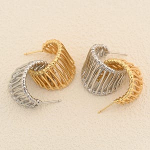 May include: Two pairs of hoop earrings. One pair is gold-toned, and the other is silver-toned. Each earring has a unique, openwork design with a curved shape. The earrings are displayed on a light beige surface.