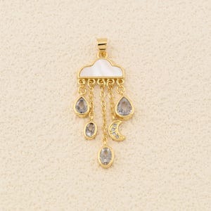 Op de afbeelding: Een goudkleurige hanger met een wolkenontwerp met een parelmoeren inleg. De hanger heeft hangende druppel- en ovaalvormige heldere stenen, samen met een halve maan bedel. De hanger is opgehangen aan een gouden ketting.
