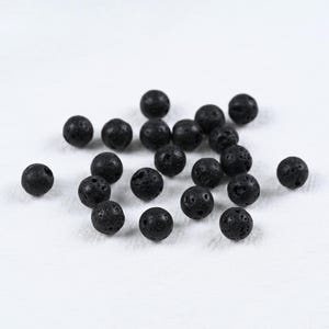 May include: A collection of small, round, black lava rock beads scattered on a white surface. The beads have a porous texture and are of varying sizes, suitable for jewelry making or aromatherapy.