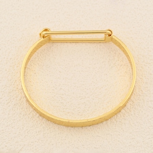 May include: A gold-colored bangle bracelet with a rectangular bar closure. The bracelet has a textured surface and a simple, elegant design. The bangle is a classic piece of jewelry.
