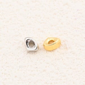 May include: Two small, metallic beads. One is silver-colored, the other is gold-colored. Both have a unique, irregular shape with a hole through the center. The beads are displayed on a textured, off-white surface.