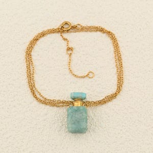 May include: A gold-colored chain necklace with a small, rectangular, light blue perfume bottle pendant. The bottle has a gold-colored cap and a textured surface. The necklace is displayed on a light-colored background.