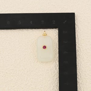 Puede incluir: Un colgante rectangular blanquecino con una peque&ntilde;a gema roja y redonda en el centro. El colgante tiene una argolla dorada en la parte superior para sujetarlo a una cadena. El colgante mide aproximadamente 2,5 cm de largo.