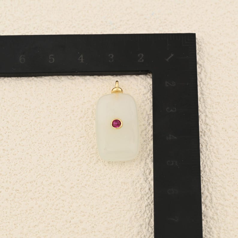 Puede incluir: Un colgante rectangular blanquecino con una peque&ntilde;a gema roja y redonda en el centro. El colgante tiene una argolla dorada en la parte superior para sujetarlo a una cadena. El colgante mide aproximadamente 2,5 cm de largo.