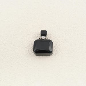 May include: A small, black, faceted perfume bottle with a rectangular shape and a matching black cap. The bottle features a metallic silver-toned collar connecting the cap to the main body, which has multiple flat, angled surfaces reflecting light.