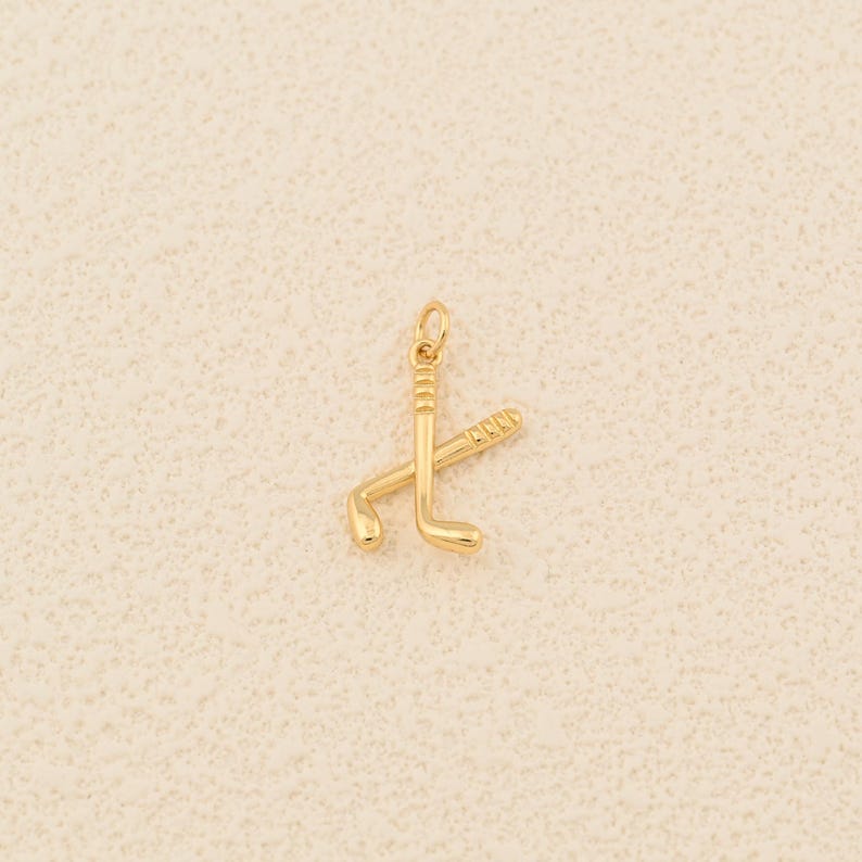 May include: A gold-colored charm featuring two crossed hockey sticks. The charm has a small loop at the top for attaching to a necklace or bracelet. The hockey sticks are detailed with grooves and curves.