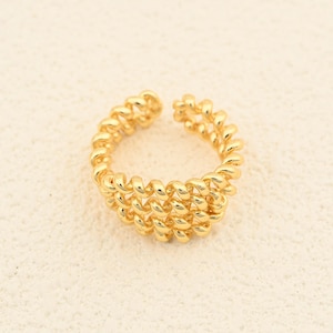 May include: A gold-colored ring with a spiral design. The ring has an open design and is made of a shiny, metallic material. The ring is shown on a cream-colored surface.