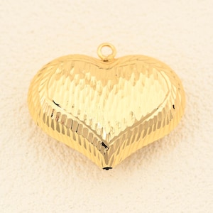 May include: A gold-colored heart-shaped pendant with a textured surface. The pendant has a small loop at the top for attaching to a chain or cord. The heart's surface has a ridged pattern.