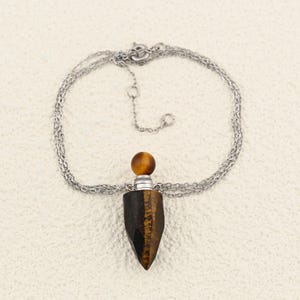 May include: A silver chain necklace with a pendant featuring a tiger's eye gemstone. The pendant is a dark brown, faceted shape with a rounded, amber-colored top. The necklace is displayed on a cream-colored surface.